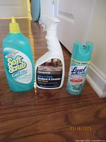 Three cleaning bottles: Soft Scrub Gel Bleach, Armstrong Resilient & Ceramic Floor Cleaner, Lysol Disinfectant Spray arranged side by side on wooden floor.