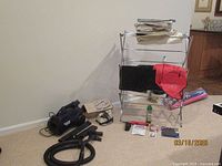 Photo showing vacuum cleaner, iron box, clothes drying rack with garment bags, and other household items on carpeted floor.