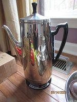 Photo of a tall, shiny metal General Electric coffee pot with a black handle and black knob on lid, electricity cord attached