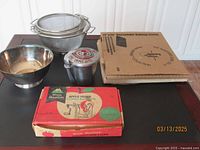 Photo shows a tabletop with the White Mountain Apple Parer Corer & Slicer box, three metal colanders, a silver plated bowl, a small ice cream server container, and two baking stones stacked beside each other.