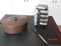 Brown LeCreuset oval Dutch oven with lid, metal spatula, and revolving spice rack on black table.