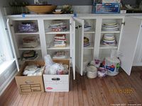 Wide shot showing cupboards open with stacks and boxes of paper plates, bowls, napkins, and packages of cups and cutlery