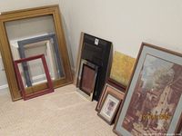 Photo showing a variety of assorted picture frames and framed art pieces leaning against a wall on a carpeted floor, highlighting the diversity in frame sizes and some included artwork such as a village scene and portrait.
