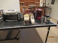 Lot of small kitchen appliances on black table including DeLonghi convection oven, Keurig coffee maker, two white Osterizer attachments, and Osterizer blender with glass pitcher