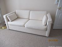 Front view of cream convertible sofa with two matching side pillows, on beige carpet next to a door and lamp.