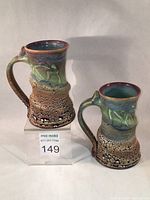Two ceramic mugs with textured snakeskin glaze and copper red trimmed rims, standing on clear block with lot number card