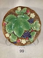 Full view of circular majolica plate showing large central grape leaf, purple grape bunches, strawberry plant leaves and flowers, all in relief with green, blue, brown, and pink glazes.