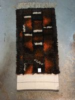 Full view of the rya rug showing dark brown, burnt orange, black, and white shaggy wool with fringed end and woven backing.