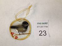 Front view of round glass ornament with chickadee perched on a branch with red berries and sheer gold hanging ribbon