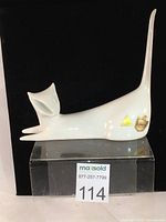 White ceramic stylized cat figurine shown from front and side angles with tall tail upright. Visible original Royal Dux pink triangle and circled triangle stamps as well as two gold and yellow stickers on the cat's side. Displayed against a black background on a clear stand with a Maxsold lot number.