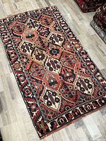 Full view of the antique Bakhtiari handmade wool rug laid flat on wood floor, showing the geometric and floral pattern in red, cream, navy, and brown.