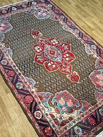 Full view of the rectangular Koliyayi handmade Kurdish Persian rug with a central red medallion and floral patterns on a brown background with navy and cream decorative borders, laid on hardwood floor.