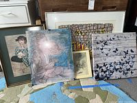 Five framed prints arranged leaning against furniture, depicting people, butterfly art, and children.