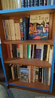 Three-shelf wooden bookcase with various books on Jewish topics including 'The Illustrated Torah' prominently displayed on the middle shelf.