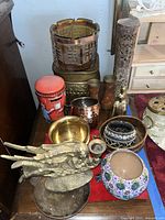Photo showing multiple small decorative items including brass candle holder angel, brass figurine bell, various bowls, copper cup, painted vases, and a red cylindrical container.