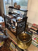 Photo showing ornate wooden shelf with scrollwork containing various decorative items including the brass handled bowl, figurines, and trays; brass crown-shaped vessel and shell-shaped bookends visible nearby among other items.