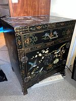 View of the front of the black lacquered cabinet showing the drawer with two handles and the double door cabinet with floral and bird painted motifs, including metal hardware and signs of wear.