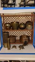 Wicker shelf filled with two copper bowls, brass vases, brass candle holder, and brass animal figurines including a turtle and frog.