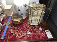 Overview of red surface featuring multiple gold-tone necklaces, a wristwatch, a small bejeweled keepsake box, and part of a brass and glass display case with miniature perfume bottles inside.