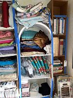 Wide view of shelving unit with multiple folded fabric pieces including quilts and bolts of fabric stacked horizontally.