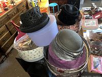 Photo showing a stack of various women's hats and hat boxes, including a black woven hat with floral cut-outs, a purple woven hat, and a gray felt hat with butterfly brooch.