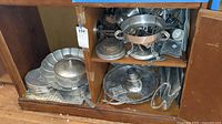 Wide view of wooden cabinet shelf holding various metal serving trays, plates, bowls, and cups with vintage designs.