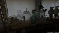 Photo showing silhouette of multiple glass jars with lids, some ornate and colored glass bottles, placed in front of bright window.