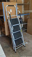 Photo of the white folding step ladder leaning against a pegboard wall, showing four wide black steps with non-slip tread and black foam hand grips.