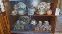 Full view of wooden cabinet shelf showing arranged tea set, plates, stemware and shot glasses
