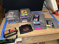 Multiple stacks of NES game cartridges with labels visible like Super Mario Bros 3, Tetris, Mario Bros, and video game accessories Game Genie and The Power Glove.