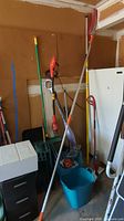 Various yard tools including Homelite electric weed wacker, shovel, branch saw, brooms, buckets, and organizer seen leaning against wall with blue bucket below