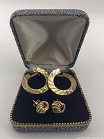 Pair of 18kt hoops and 10kt knotted studs displayed side by side in a hinged jewelry box with velvet-lined interior