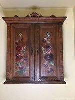 Front view of wooden antique apothecary medicine cabinet with hand painted floral panels and ornate carved wood details on sides and top molding.