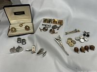 Overview of entire cufflink and accessory assortment on white fabric