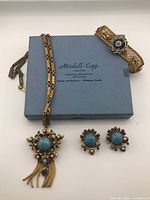 Photo showing the full set including necklace with large turquoise faux stone pendant and chain tassels, matching clip-on earrings, and gold-toned mesh bracelet with pearl floral medallions on blue enamel.