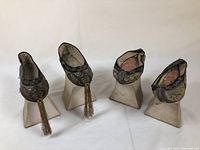 Front view of two pairs of Manchu Chinese platform shoes on white stands showing detailed embroidery and tassels on the black pair and rolled fabric tops on the multicolored pair.