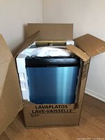 Front view of dishwasher inside open cardboard box, blue protective film on door, foam inserts in place