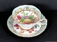 Front angle showing teacup sitting on saucer displaying fruit design and gold floral pattern on blue background