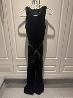 Full view of black velvet sleeveless jumpsuit hanging on a hanger from top to bottom showing entire outfit and satin belt tied at waist.