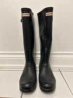 Front view of pair of black Hunter rain boots, showing tall rubber design with logo badge and rounded toe.