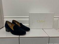 Pair of navy blue velvet heeled loafers displayed next to a white Chloé branded box, front perspective.
