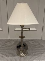 Full view of brushed nickel table lamp with white fabric shade on, placed on carpet in front of closed doors.