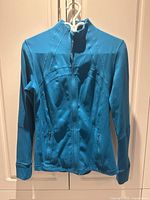 Front view of teal Lululemon women's yoga jacket size 10 hanging on white hanger against cabinet doors.