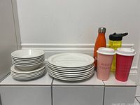 Photo showing stacks of white ceramic Mikasa dishes including plates and bowls with four Starbucks reusable cups and two water bottles in back.