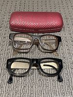 Two pairs of prescription eyeglass frames laid flat with red textured eyeglass case above them on fabric surface