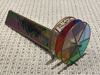 Side angled view of the kaleidoscope showing the iridescent prism-shaped body and colorful stained glass circular rotating section with dried flowers inside.