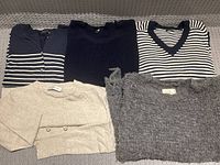 Five women's knit tops and sweaters laid out flat on a carpeted floor, showing colors and different textures.