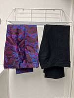 Two pairs of trousers hanging on a rack: one floral patterned in purple, blue, raspberry colors, and one black flared pair.