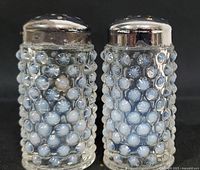 Front view of vintage opalescent hobnail salt and pepper shakers with silver tops on black background.