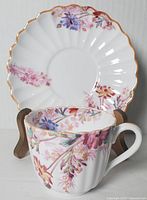 Front view of cup with saucer behind, showing floral pattern and gold trim on both pieces.